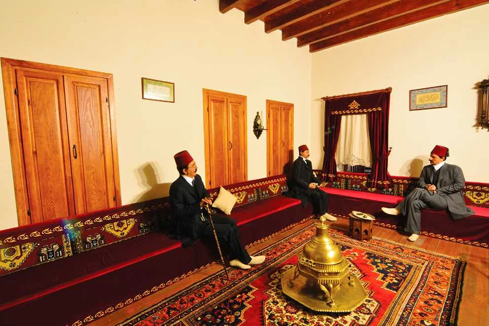 Kuruköprü Anıt Müzesi ve Geleneksel Adana Evi Geleneksel Türk oturma odası, Osmanlı kıyafetli üç adam sohbet ediyor, sıcak ve davetkar atmosfer.