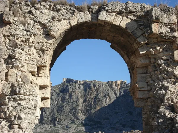 Eski taşlardan oluşan kemerin arkasında tarihi bir kalenin silueti ve mavi gökyüzü görünüyor.