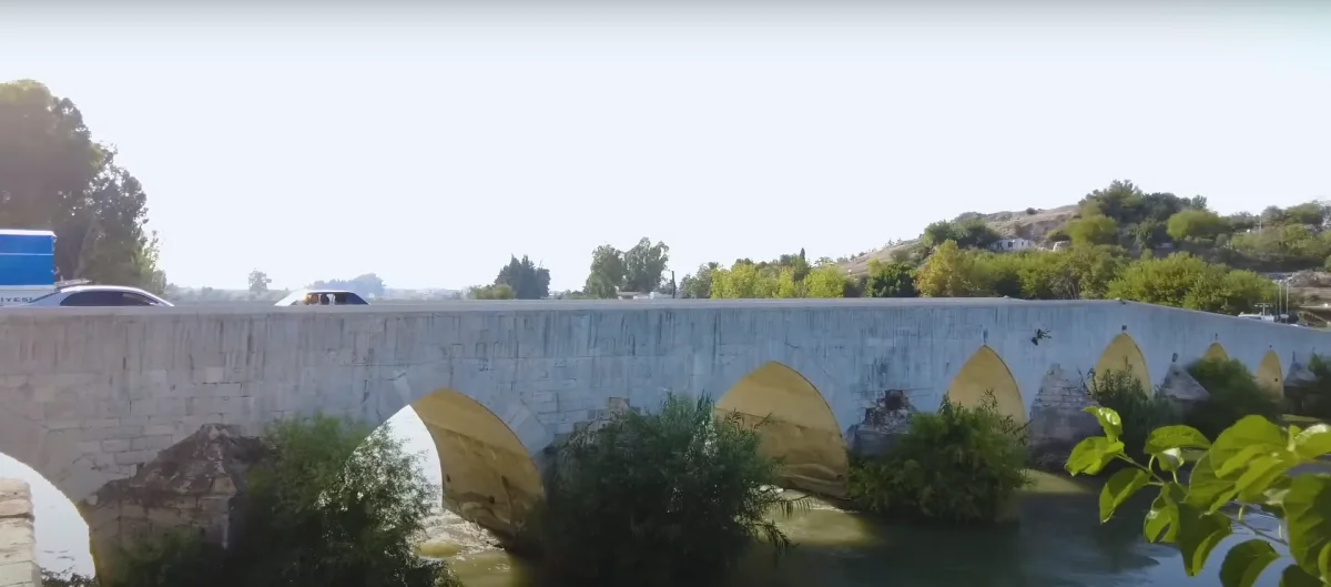 Taş yapılı köprü, yeşil ağaçlarla çevrili su üzerinde uzanıyor.
