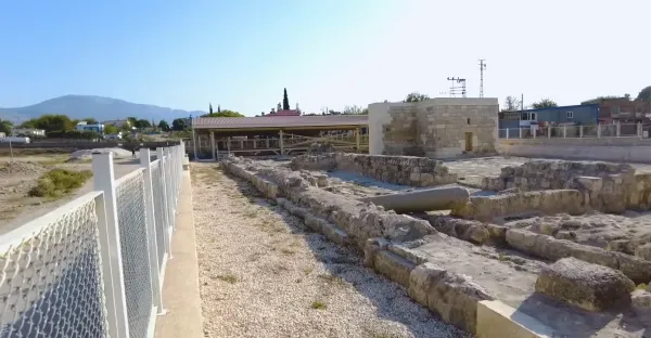 Antik kalıntılar, modern yapı ve dağlarla çevrili arkeolojik alan görüntüsü.