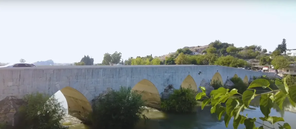 Geniş taş köprü, yuvarlak kemerler ve akıntılı nehir manzarasıyla doğal güzellik sunuyor.