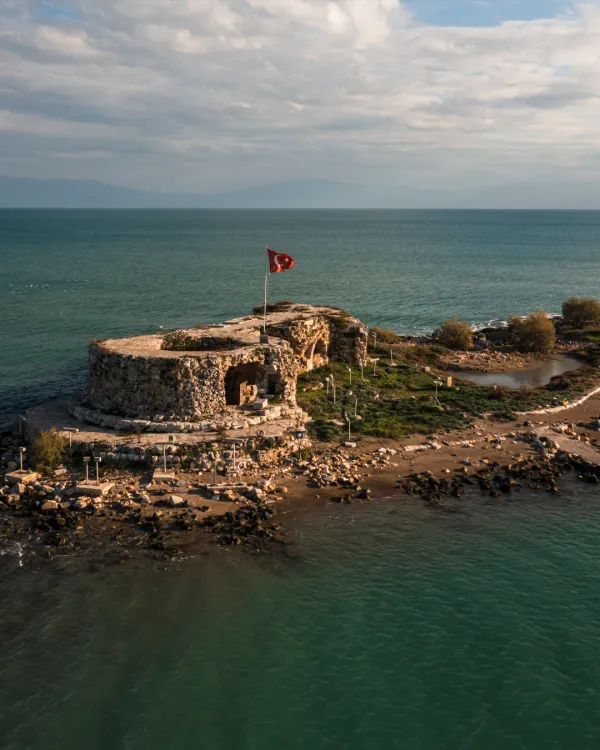 Denizle çevrili tarihi kalıntı, Türk bayrağı, yeşil bitki örtüsü ve mavi-yeşil su.