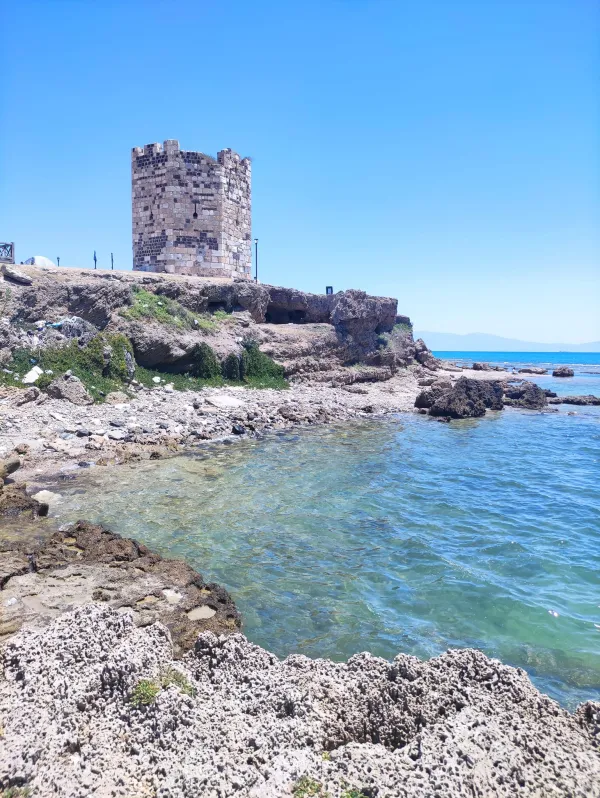 Deniz kenarında yer alan tarihi taş kule, mavi gökyüzü altında doğal bir manzara oluşturuyor.