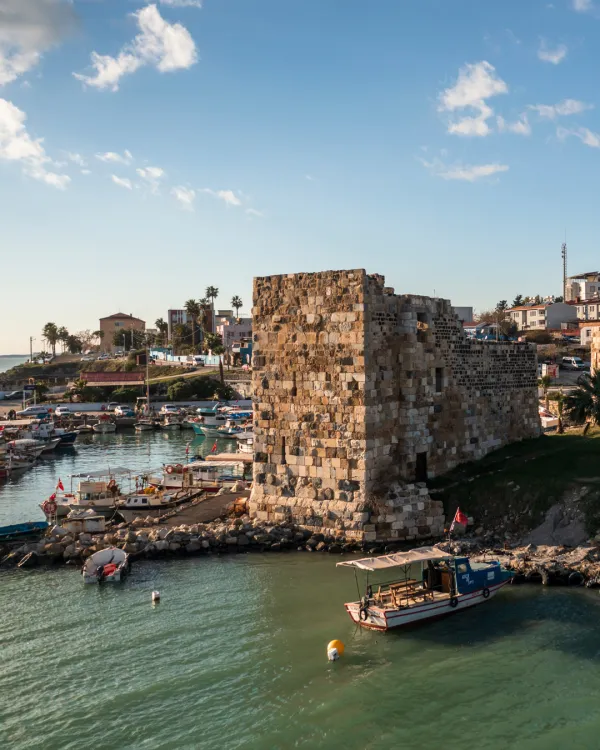 Tarihi taş surların yanında turkuaz su ve tekneler, palmiyeli modern binalar.