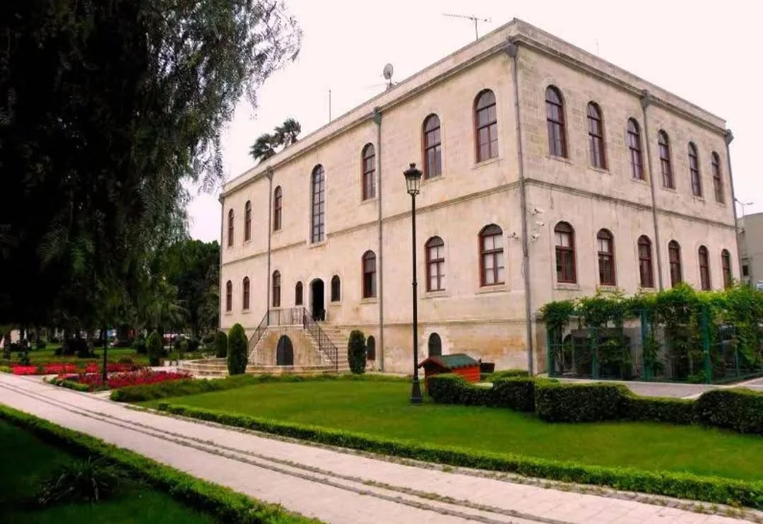 Adana Tarihi Kız Lisesi: Mimari Güzellik Büyük bir tarihi bina yeşil alanlarla çevrili, geniş pencereleri ve merdivenli giriş kapısıyla dikkat çekiyor.