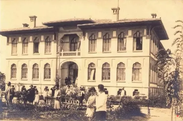 Tarihi bina, yuvarlak kemerli kapı ve büyük pencerelerle çevrili, bahçede insanlar neşeli bir buluşma yapıyor.