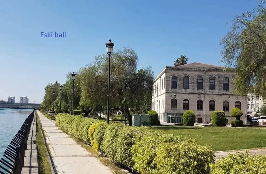 Adana Tarihi Kız Lisesi Parkı'nda Huzur Verici Manzara Yeşil park alanında yürüyüş yolu ve tarihi bina, mavi gökyüzüyle huzurlu bir atmosfer sunuyor.