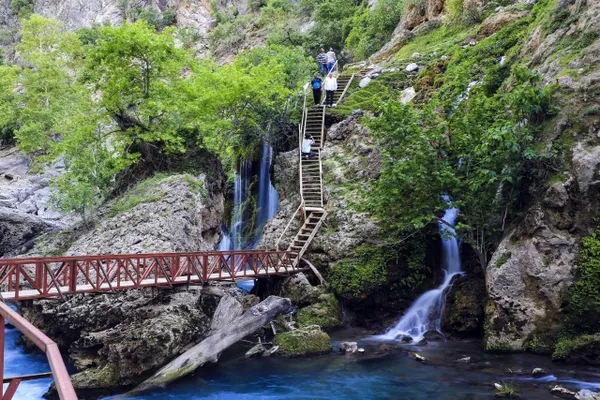 Küp şelaleleri, yeşil bitki örtüsü ve tahta köprü ile doğal manzara.