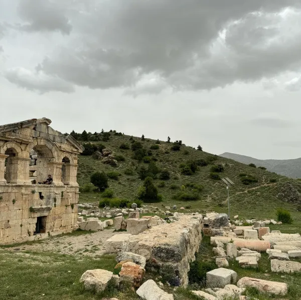 Antik yapı kalıntıları önünde oturan bir kişi ve doğal manzara.