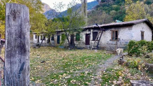Taş evin önünde dökülmüş yapraklarla kaplı bahçe ve dağ manzarası.