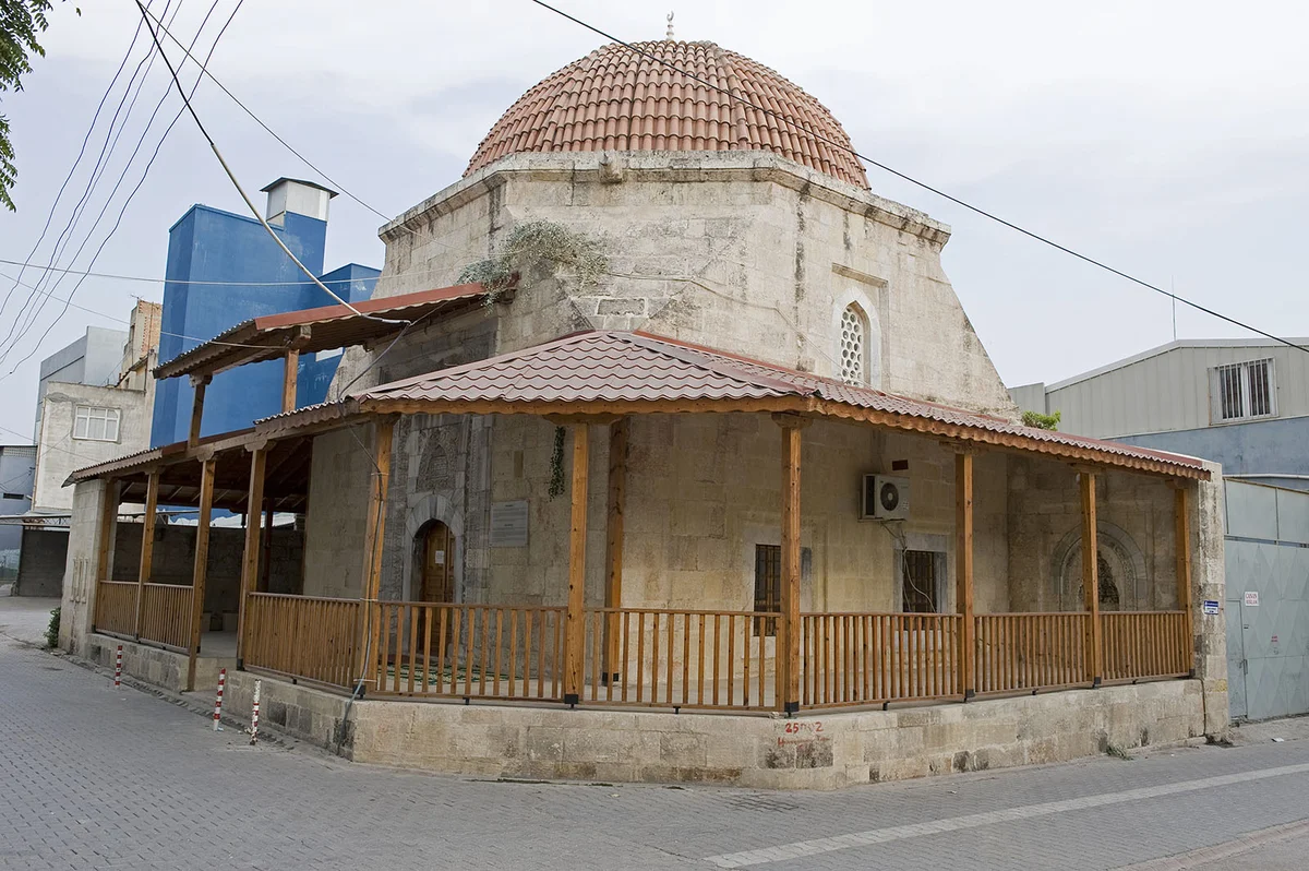 Taş yapılı tarihi Hasan Ağa Camii, ahşap destekli geniş veranda ve modern binaların arka planda yer aldığı bir görüntü.