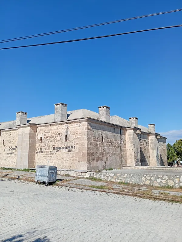 Taş duvarlardan yapılmış tarihi Kurtkulağı Kervansarayı, mavi gökyüzü altında yeşil alanlarla çevrili.