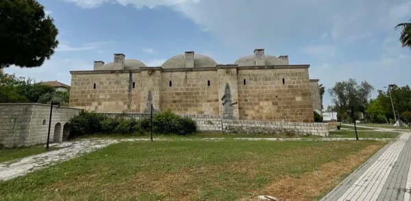 Büyük taş yapı, yuvarlak kubbeler, açık renkli duvarlar ve yeşil alan.