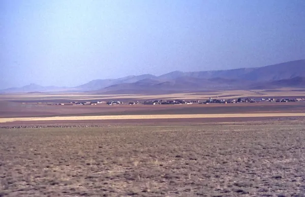 Geniş arazideki sarı, kahverengi ve yeşil tarlalar, uzak dağlar ve yerleşim yeri.