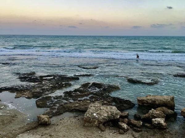 Açık mavi denizde dalgalar sahile vurmakta, arka planda bir kişi suda yürümekte.