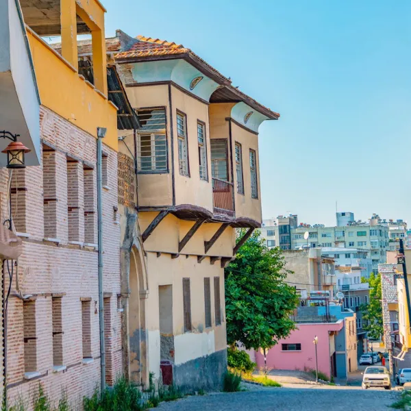 Dar sokakta yer alan tarihi balkonlu binalar, modern yapılar ve yeşil ağaçlarla çevrili.