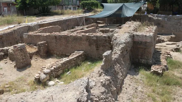 Arkeolojik alanda tuğla duvarlar, tente ve yeşil bitki örtüsü görünmekte.