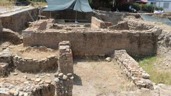 Arkeolojik kazı alanında tuğla ve taş kalıntıları, gerilmiş tente ve ağaçlar görünmekte.