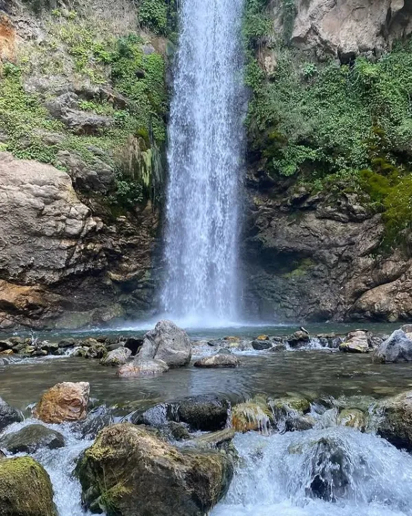 Yüksek kayalıklardan dökülen suyla çevrili yeşil alan ve gölet manzarası