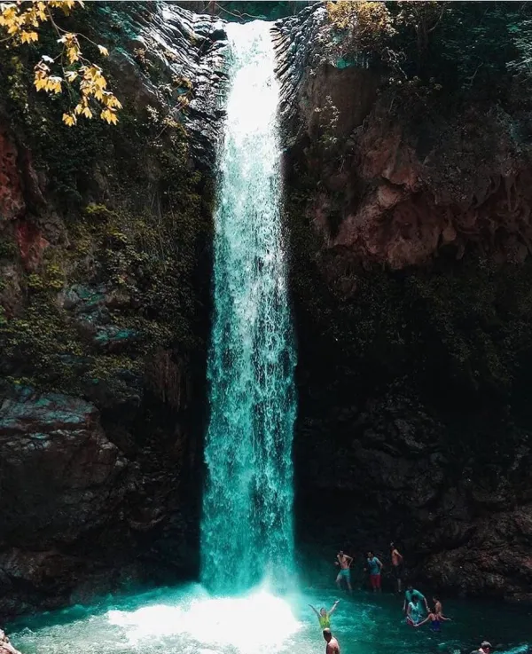 Yüksek bir şelaleden dökülen su, turkuaz havuzda insanlarla dolu yemyeşil doğada akıyor.