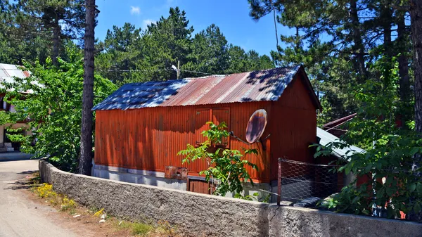 Kırmızı metal çatılı ev, çevresinde yeşil ağaçlar ve taş duvarla huzurlu doğal ortam.