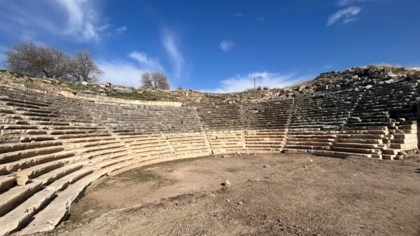 Taş oturma sıralarıyla çevrili antik tiyatro alanı, açık mavi gökyüzü ve ağaçlarla çevrilidir.