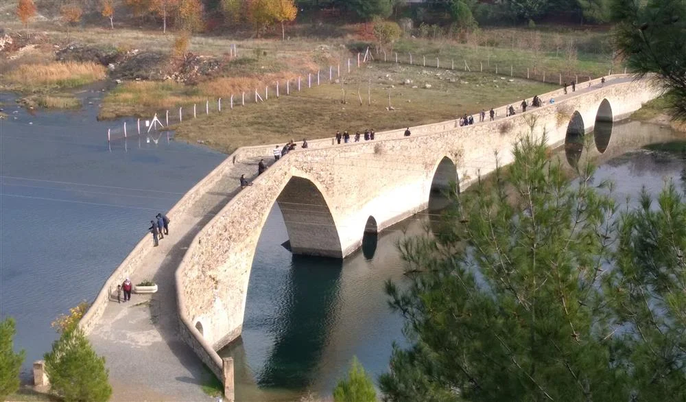 Taş köprüde yürüyen insanlar, su ve sonbahar renkleriyle çevrili doğal manzara.