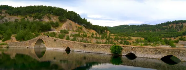Taş köprü, su üzerindeki yansıması ve yeşil dağlar arka planda.