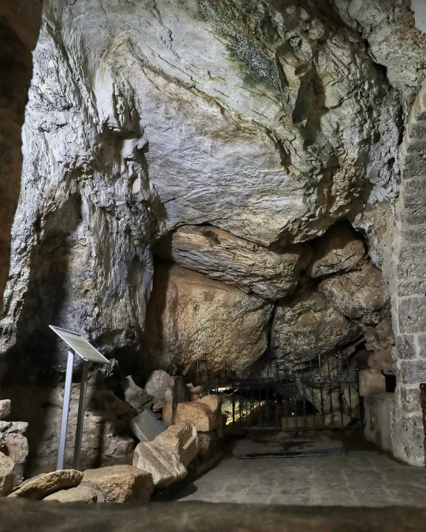 Büyükçe bir mağara, kayalık duvarlar ve bilgi panosu ile karanlık bir atmosfer sunuyor.