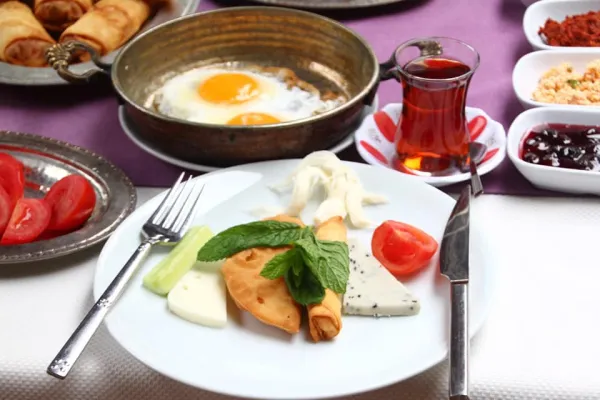 Zengin bir kahvaltı platosu, mavi-beyaz tabakta taze domates, salatalık, peynir ve börekler var.