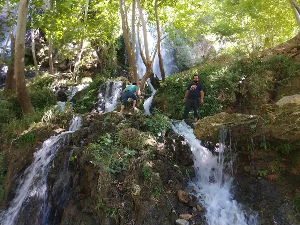 Yeşil ağaçlarla çevrili şelale ve kayalık alanda iki kişi