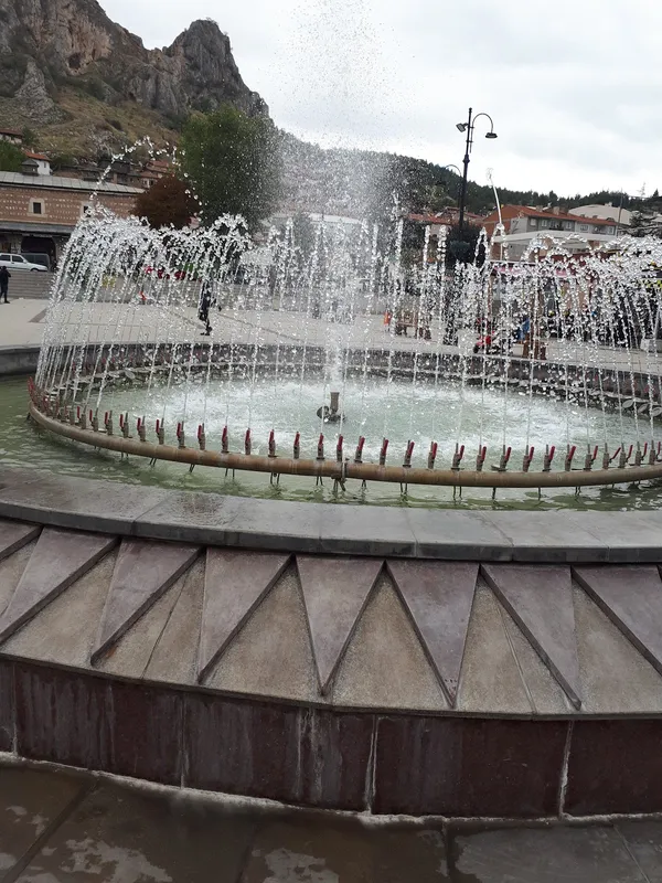 Yuvarlak havuzdan su fıskiyeleri yükseliyor, dağlık manzara arka planda görünüyor.