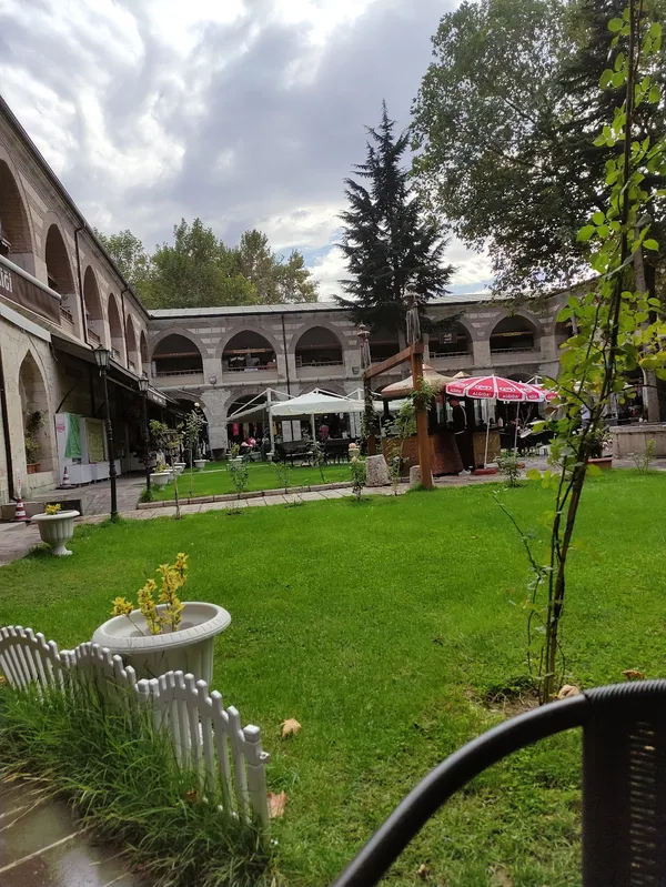 Yeşil alanda tarihi taş han, kemerli dükkanlar ve bahçe düzeniyle dinlendirici bir ortam sunuyor.