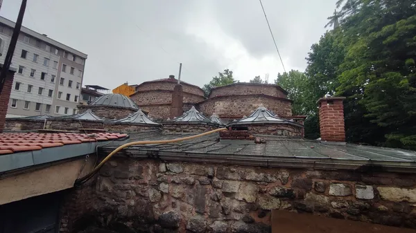 Tarihi taş medrese çatısı ve arka planda yuvarlak kubbeler ile yeşil ağaçlar