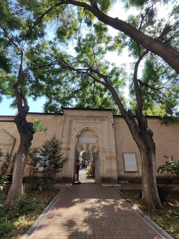 Yeşil ağaçların gölgesinde ilerleyen taş döşeli yol ve kemerli kapı.