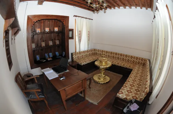 Dedeoğlu Konağı'nda ahşap kirişli oturma odası, lamba ve kitap rafı ile sıcak bir atmosfer sunuyor.