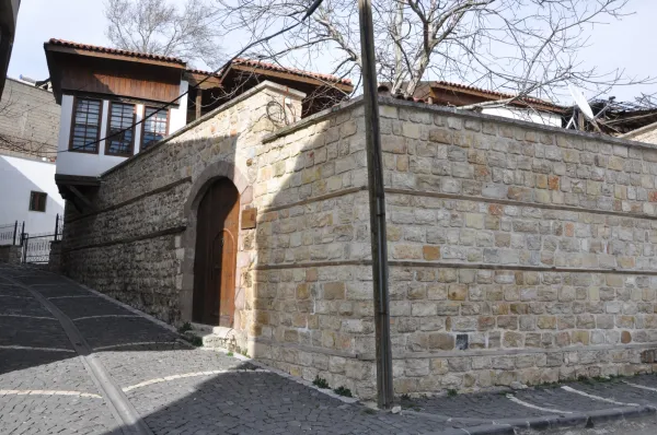 Taş duvar önünde eğimli sokak, yuvarlak kapı ve ahşap detaylar.