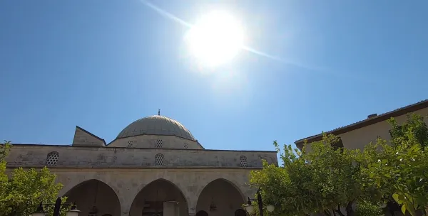 Açık mavi gökyüzü altında güneşle aydınlatılan Habibi Neccar Camisi, çevresinde doğal bir ortam sunuyor.