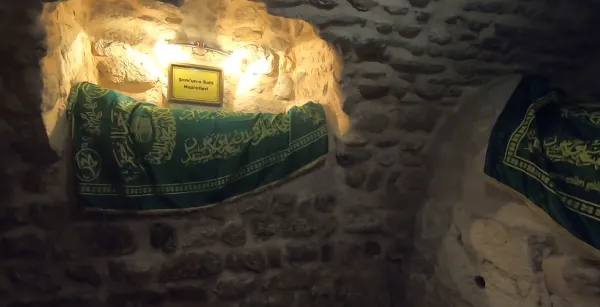 Taş duvarlarla çevrili nişte yer alan yeşil örtü ve sarı tabela.