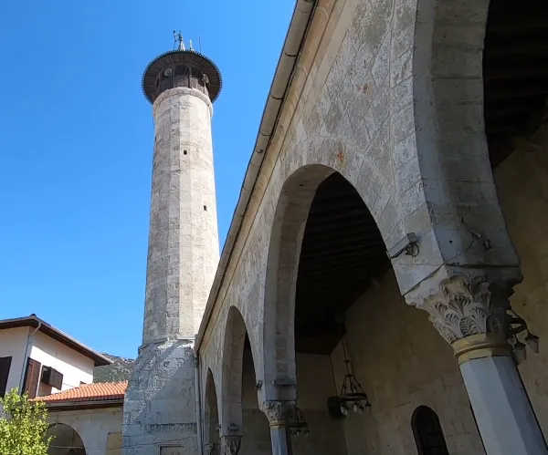 Taş kule, kemerli sütunlar ve dağlık arka planın olduğu tarihi cami görüntüsü.