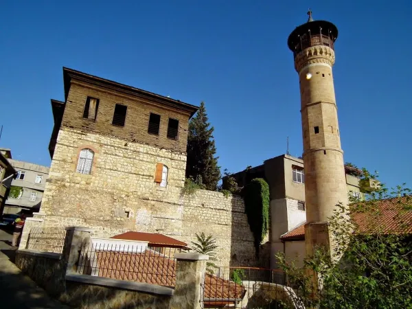 Taş yapılı iki katlı bina ve silindirik minare, yeşil ağaçlarla çevrili.