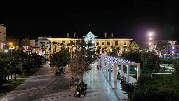Hatuniye Camii'nin aydınlatılmış yapısı, palmiyelerle dolu yürüyüş yolu ve hareketli dükkanlar.
