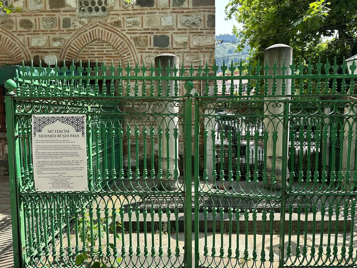 Tarih ve Huzur - Hatuniye Camii Mezarlığı, Geleneksel Taş Yapılarla Yeşil demir parmaklıklarla çevrili mezar alanı ve arka planda geleneksel taş yapı.