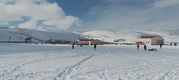 Karla kaplı alanda yürüyen ve karda oynayan insanlar, arka planda dağlar ve teleferik hattı.