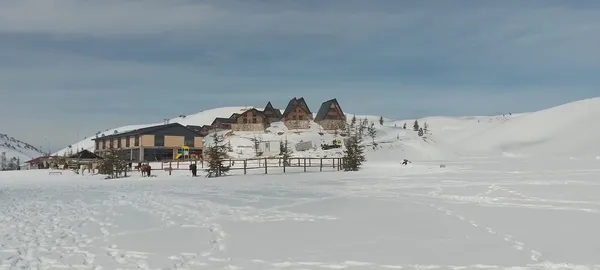 Karla kaplı geniş alan, dağlar ve tipik dağ evi mimarisiyle çevrili.