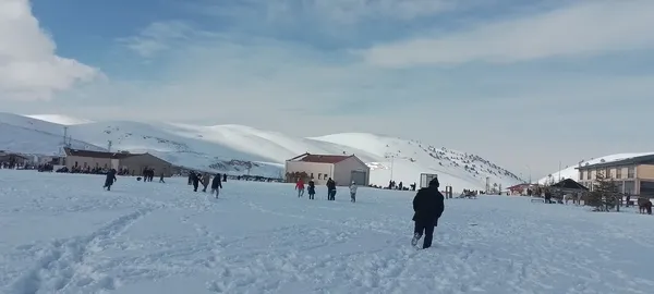 Karla kaplı alanda yürüyen ve etkinlik yapan kalabalık bir grup insan, arka planda beyaz dağlar.