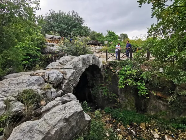 Yoğun yeşilliklerle çevrili eski taş köprüde iki kişi konuşuyor.