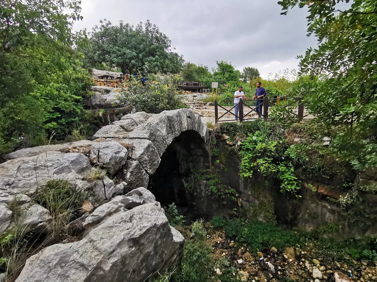 Sakin Doğanın İçindeki Eski Taş Köprü - Gizemli Bir Mağara Keşfi mi? Yoğun yeşilliklerle çevrili eski taş köprüde iki kişi konuşuyor.