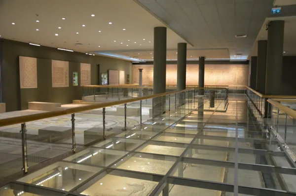 Modern müze iç mekanında yüksek tavan ve cam zeminle sergilenen antik mozaikler ve taş yapıları.