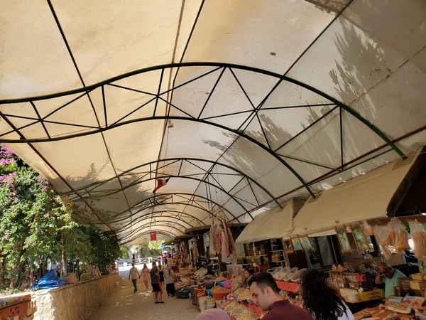 Eğri hatlarla desteklenmiş tente altında tezgahlar ve alışveriş yapan insanlar.